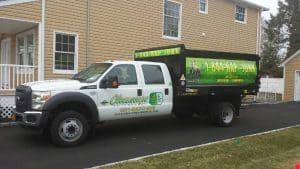 A white pickup truck with a large black dumpster bed is parked on a driveway in front of a beige house. Emblazoned with its logo and contact number, this junk removal service is ready to tackle your clutter needs efficiently across NY, including Long Island, NY.