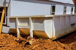 A large white metal dumpster placed beside a white-sided building with multiple windows, surrounded by reddish-brown dirt and debris at a construction site in Long Island, NY.