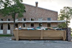 A large rectangular dumpster filled with debris sits in front of a brick building with multiple windows and doors, surrounded by trees in a parking lot area in Long Island, NY.