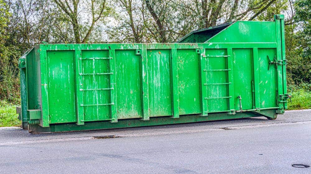 A large green dumpster sits on the side of a paved road, with trees and greenery in the background. The dumpster shows some signs of wear and rust.