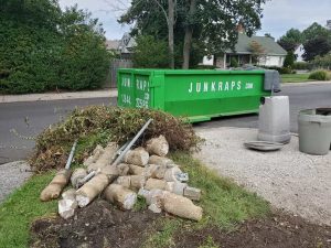 A pile of uprooted plants and broken concrete pieces lies beside a green dumpster labeled “JUNKRAPS.COM” on a residential street. Trash bins and a house are visible in the background.